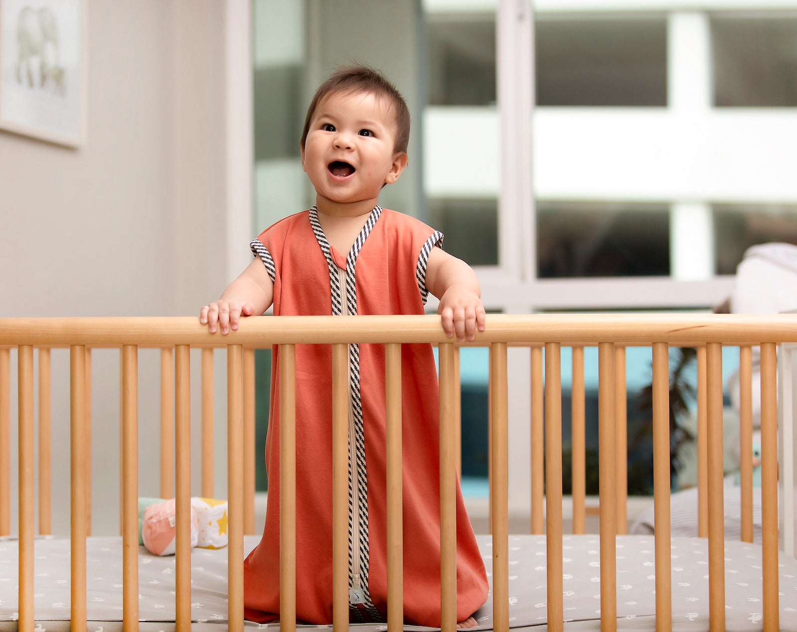 Baby standing in crib wearing a coral colored Sleeper Bag
