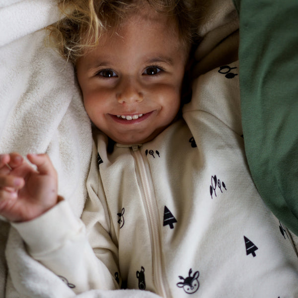Toddler smiling at the camera wearing a black forest printed footie and snuggled in a green and natural blanket
