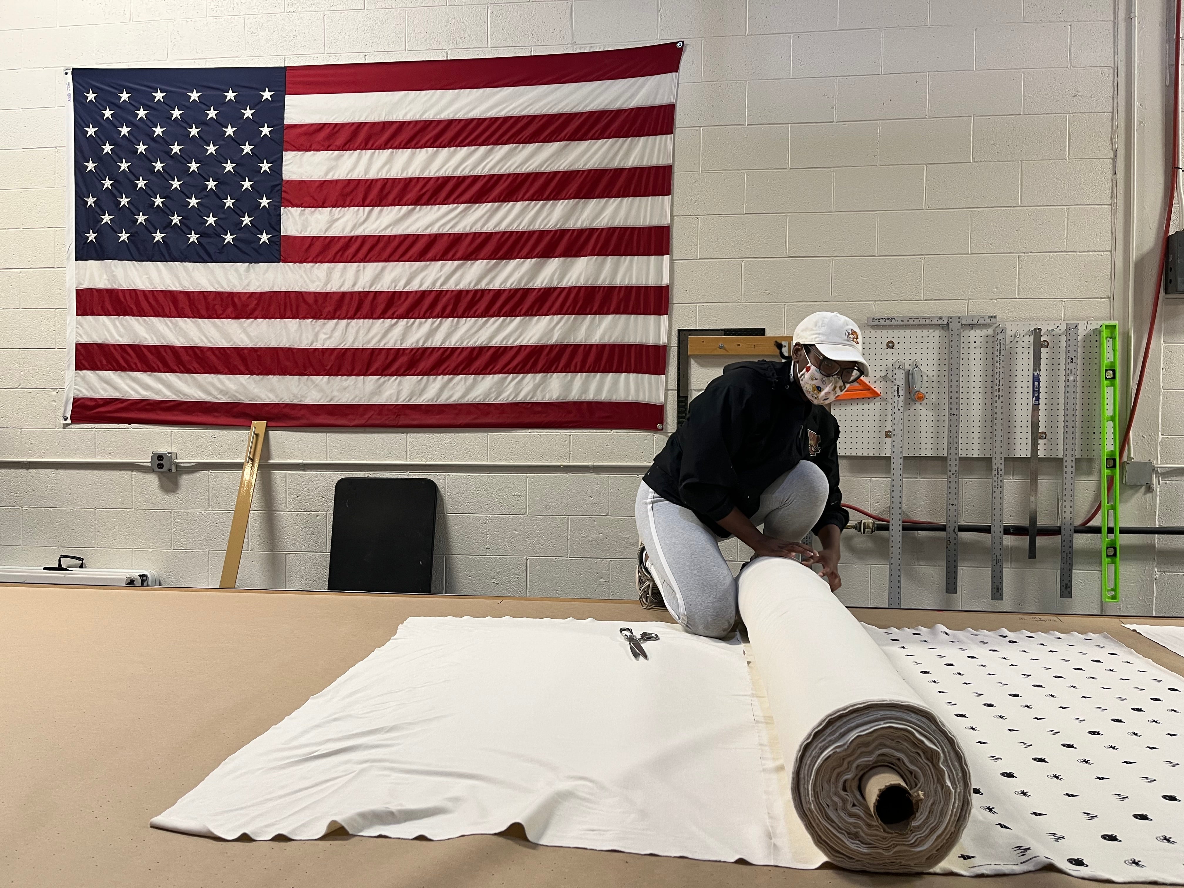Young woman spreading fabric on cutting table with American flag behind her.